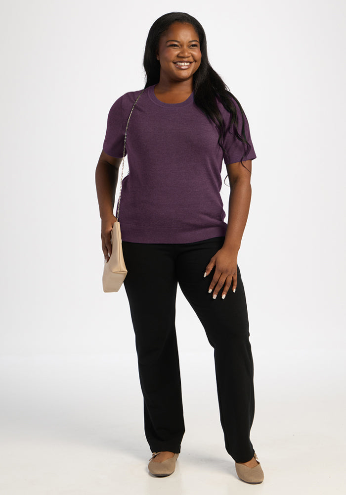 Woman smiling in Woolx Anna Crew Sweater - Purple Iris, black pants, beige shoes, holding a beige purse.