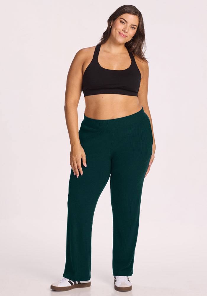 Woman in a black sports bra and Woolx Luca Ribbed Pants - Deep Pine smiles against a plain light background.