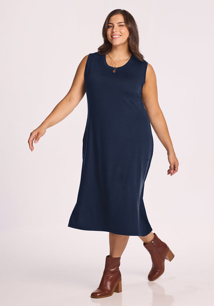 A woman wearing the Woolx Cassie Dress in Deep Navy and brown ankle boots stands against a light background, smiling as she walks forward.