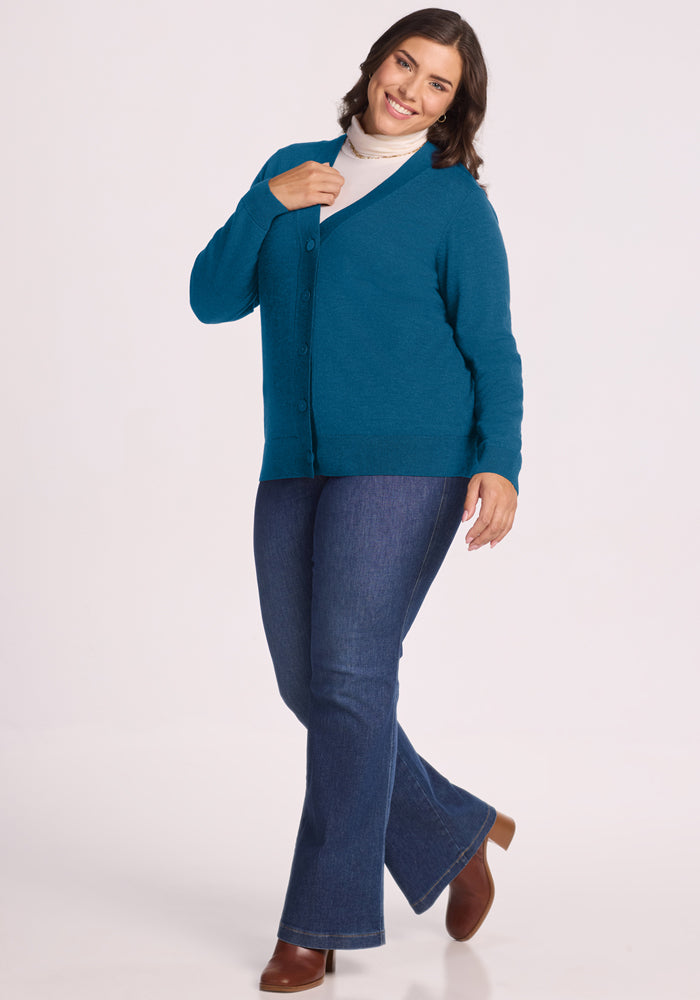 Woman in Woolx Gia Sweater - Blue Pine, white turtleneck, jeans, and boots, smiling against a white background.