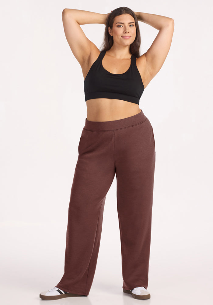 Woman in black sports bra and Woolx Bree Lounge Pants - Canyon Clay stands smiling with arms raised against white background.