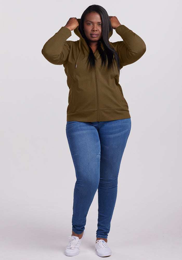 Woman in Woolx Ryann Hoodie - Olive Leaf, blue jeans, and white shoes faces camera, holding her hood.