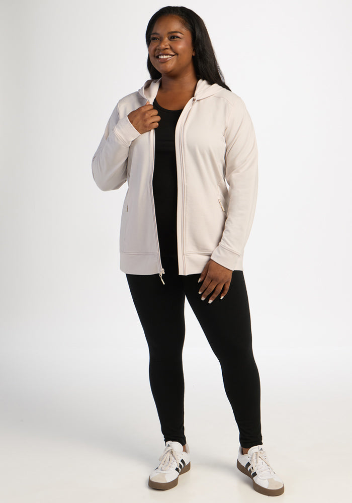 Smiling woman in Woolx Darby Zip Up Hoodie - Ecru, black leggings, and white sneakers stands against a white background.