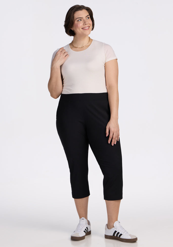 A woman with short brown hair smiles as she stands against a white background, wearing Woolx Nicole Crop Pants in black, a light beige t-shirt, and white sneakers with black stripes.