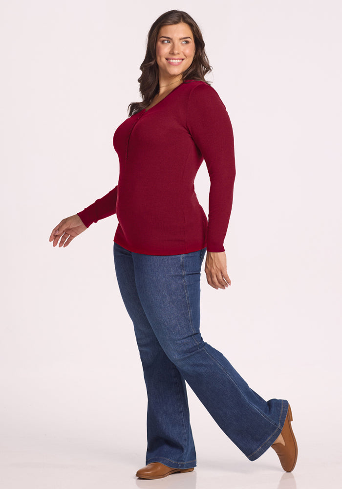 Woman in Woolx Reese Ribbed Henley - Beet Red, blue jeans, and brown shoes smiles walking sideways on white background.