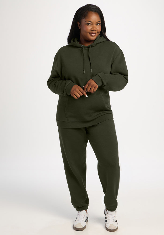 Woman in a Woolx Avery Hoodie - Duffel Bag and sweatpants stands smiling, wearing white sneakers on a plain background.