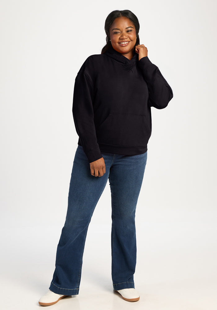 Woman in a Woolx Lizzy Hoodie - Black smiles, touching her hair against a white background, wearing blue jeans.