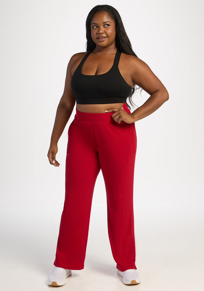 Woman in Luca Ribbed Pants - Maraschino Cherry by Woolx, black sports bra, and white sneakers stands confidently.