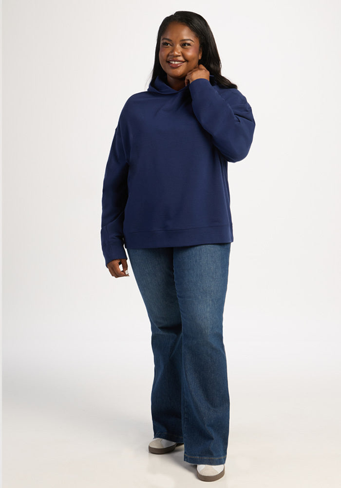 A woman wears a navy blue Woolx Genevieve Hoodie in Ocean Cavern and blue jeans.