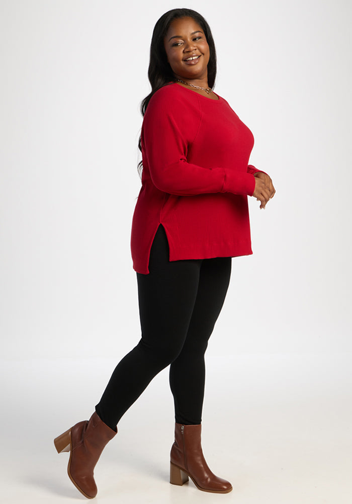 Woman in Woolx Ainsley Sweater - Maraschino Cherry, black leggings, and brown boots stands by a white backdrop.