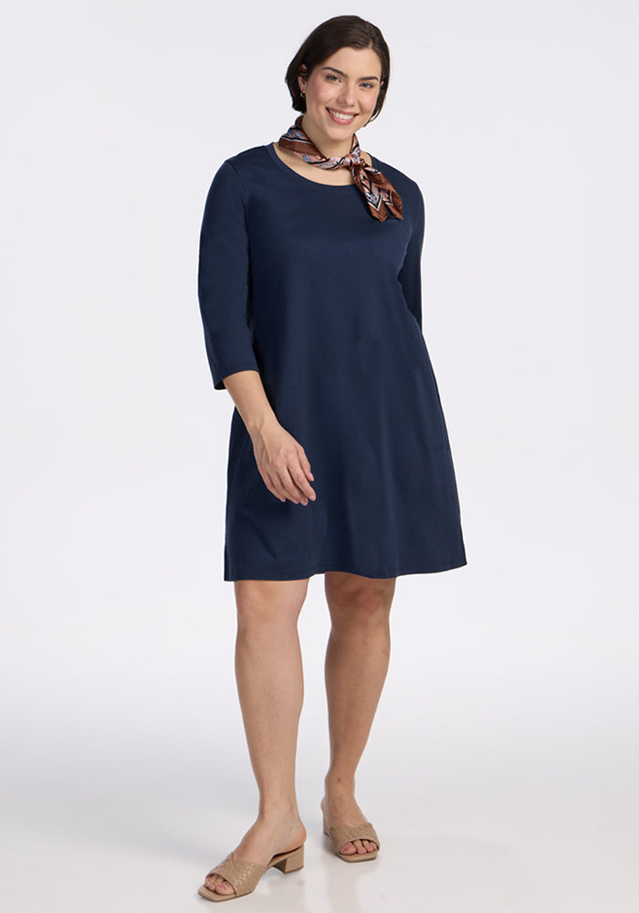 A woman models the Woolx Rissa A Line Dress in Deep Navy—knee-length Merino wool with three-quarter sleeves—styled with a patterned scarf and beige heeled sandals, ideal for fall/winter layering or as a chic travel dress. 