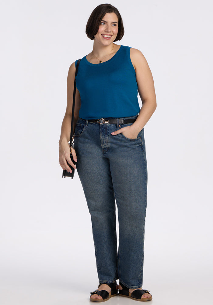 A woman with short brown hair smiles as she stands against a white background. She wears the Woolx Chessy Tank Top in Moroccan Blue, blue jeans with a belt, black sandals, and carries a small black shoulder bag. One hand is in her pocket.