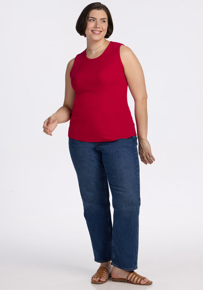 A woman with short dark hair smiles while standing against a white background, wearing the Woolx Raegan Ribbed Tank Top in Poppy Spark, blue jeans, and brown sandals.