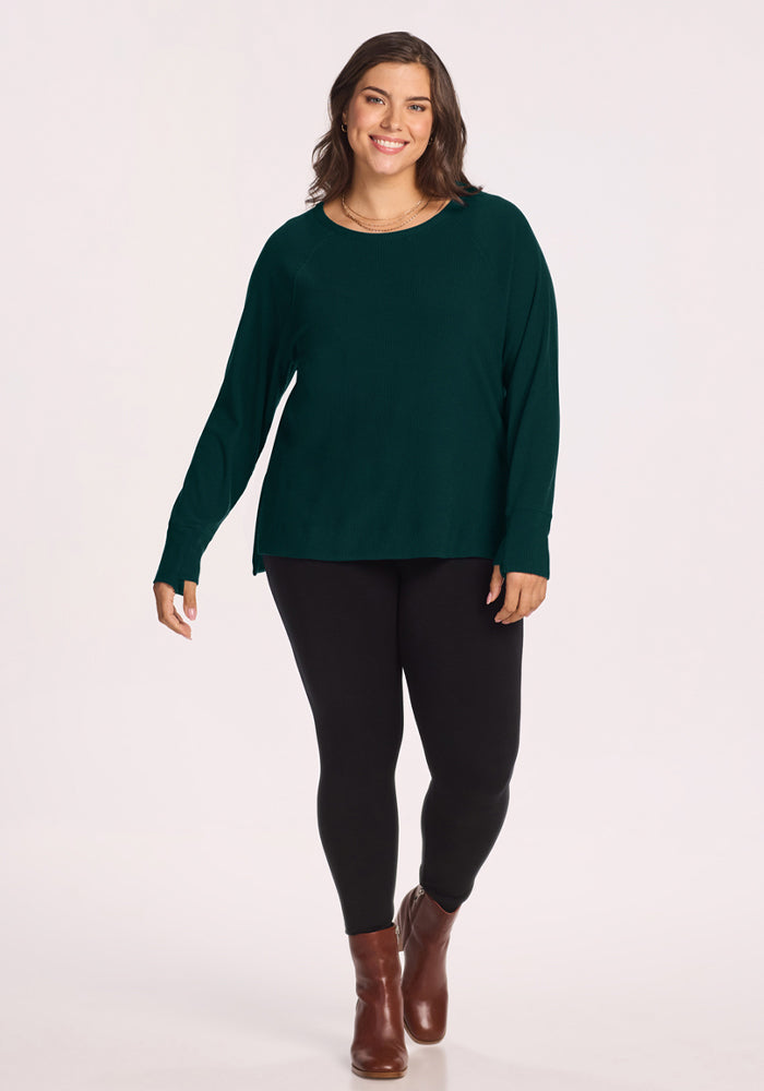 Woman in Woolx Ainsley Sweater - Deep Pine, black leggings, brown boots, smiling against a light background.