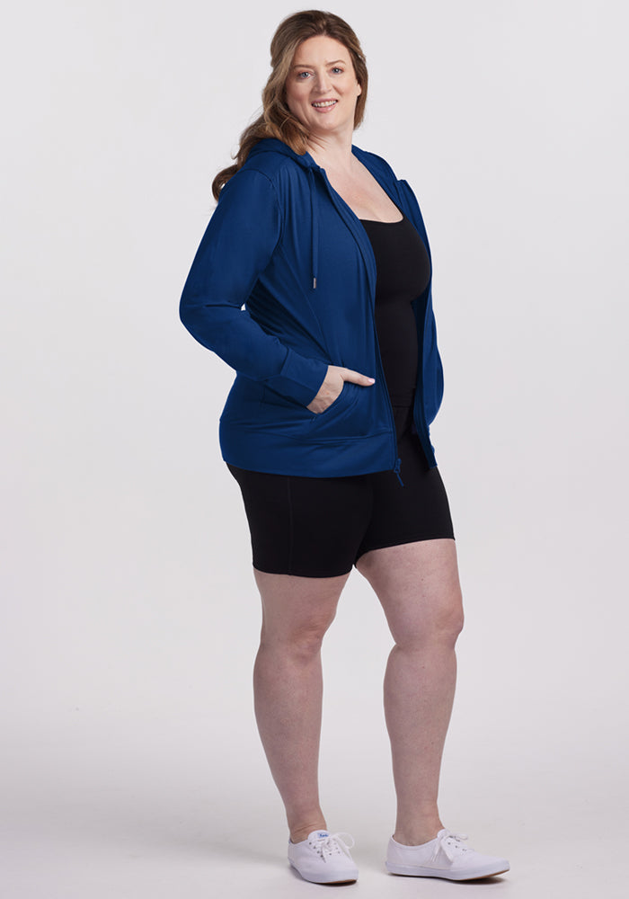 A woman with long brown hair, wearing the Woolx Ryann Hoodie in Estate Blue, black shorts, and white sneakers stands smiling against a plain white background.