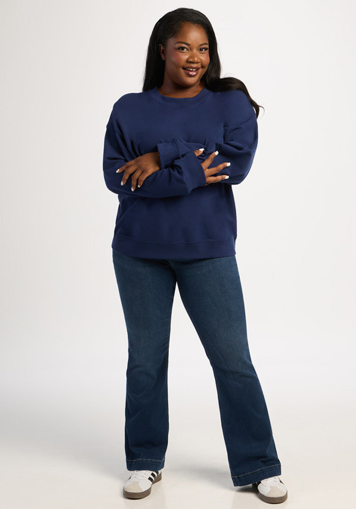 A woman wearing a Molly Crewneck - Ocean Cavern merino wool sweater and blue jeans with her arms crossed.