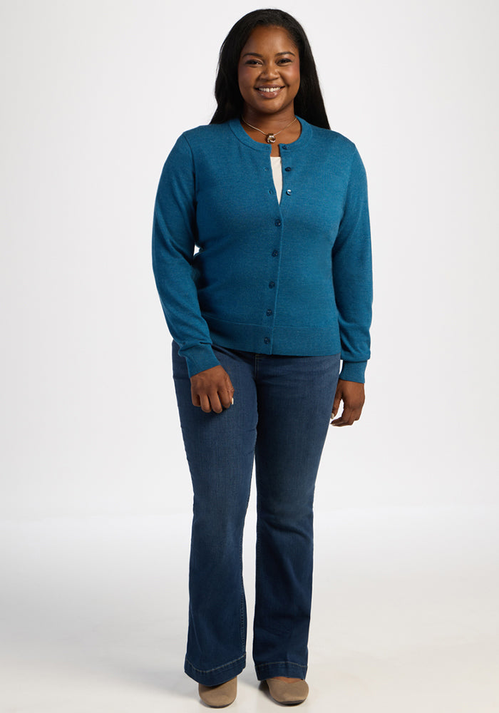 A woman in a Woolx Caitlin Sweater - Blue Pine and jeans smiles against a plain white background.