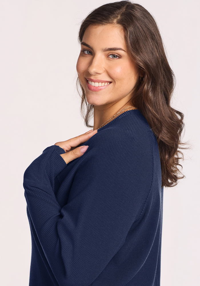 Woman with wavy brown hair smiles over her shoulder in the Woolx Ainsley Sweater—Ocean Cavern.