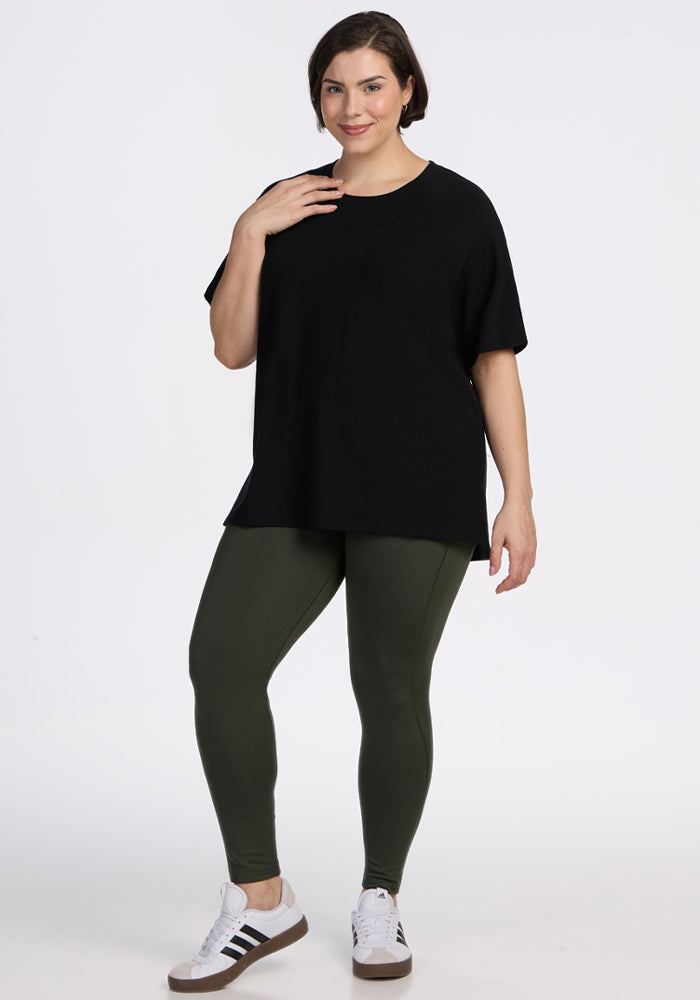 A woman stands smiling against a plain white background, wearing the Woolx Audrey Tee in black, olive green leggings, and white Adidas sneakers with black stripes.