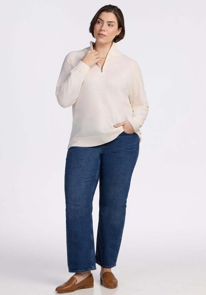 A woman with short brown hair models the Woolx Collins Half Zip Sweater in Dewdrop Pearl, paired with blue jeans and brown loafers. She stands against a plain white background, one hand in her pocket and the other touching her collar.