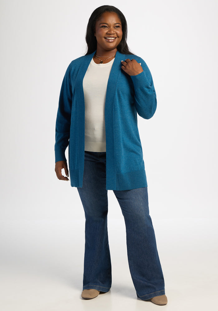 Woman in a Woolx Macie Sweater - Blue Pine, white top, and jeans stands smiling against a plain white background.