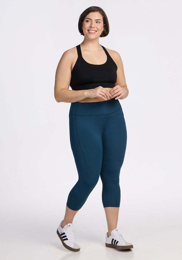 A woman smiles against a white background wearing a black sports bra, Woolx Merino Flex® McKenna Capris in Majolica Blue, and white sneakers with black stripes.