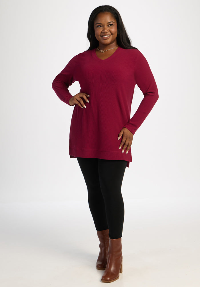 Woman smiles in a Woolx Evie Ribbed Tunic in Beet Red, black leggings, and brown boots against a plain background.