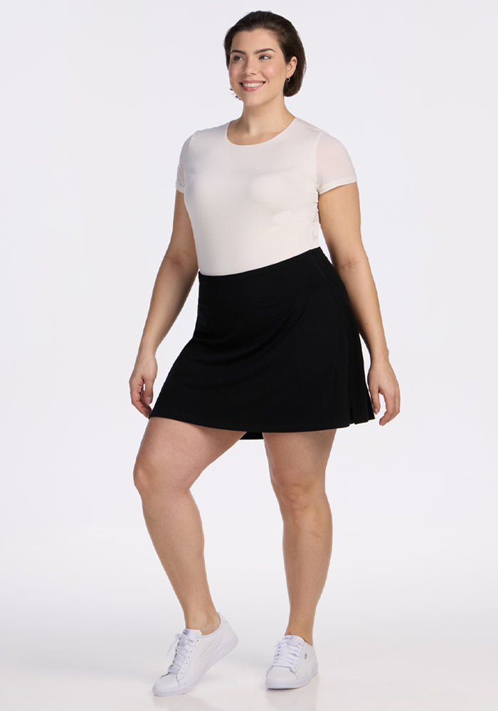 A woman stands smiling against a plain white background, wearing a short-sleeved white top, the Woolx Sienna Skort - Black with pockets, and white sneakers. 