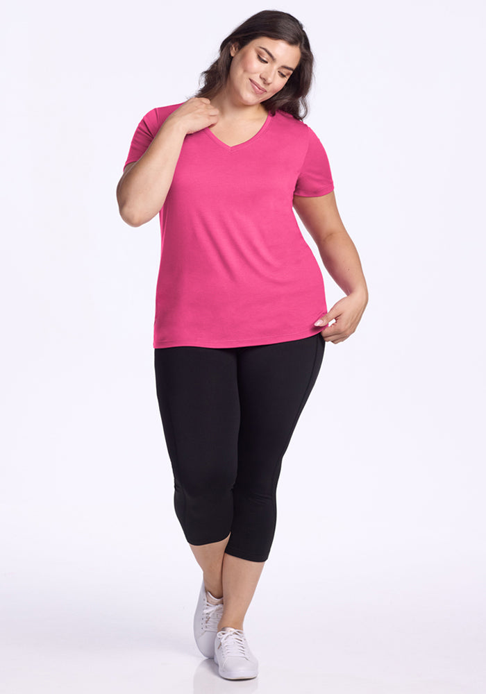 A woman smiles, wearing the Woolx Junie V Neck in Raspberry Fizz—a bright pink relaxed fit tee—paired with black capri leggings and white sneakers, posing against a plain white background.