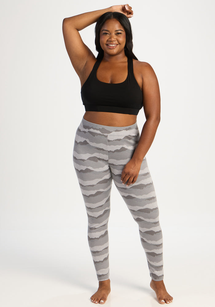 Woman in Woolx Taylor Baselayer Leggings - High Peaks, black sports bra, barefoot, smiling against white background.