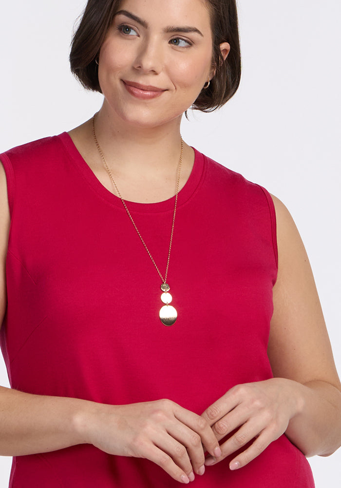 A woman in the Woolx Cassie Dress - Poppy Spark, a sleeveless, bright pink Merino wool tank dress, smiles softly as she looks to the side with her hands gently clasped in front of her, wearing a gold pendant necklace.