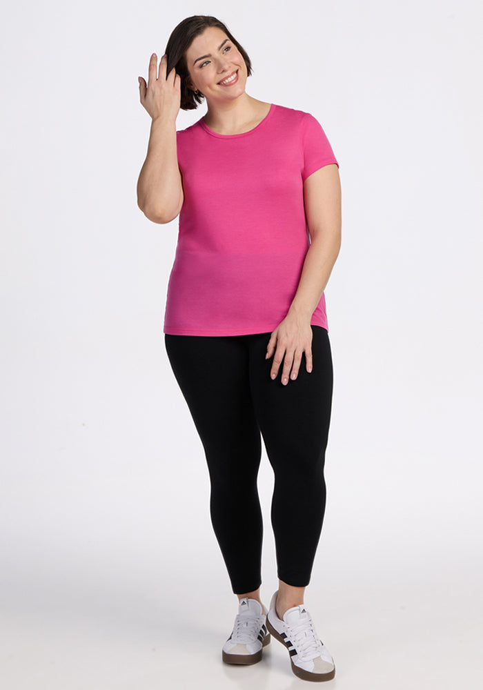 A woman wearing the Woolx Addie Short Sleeve Crew in Raspberry Fizz, black leggings, and white sneakers stands smiling and touching her hair against a plain white background.