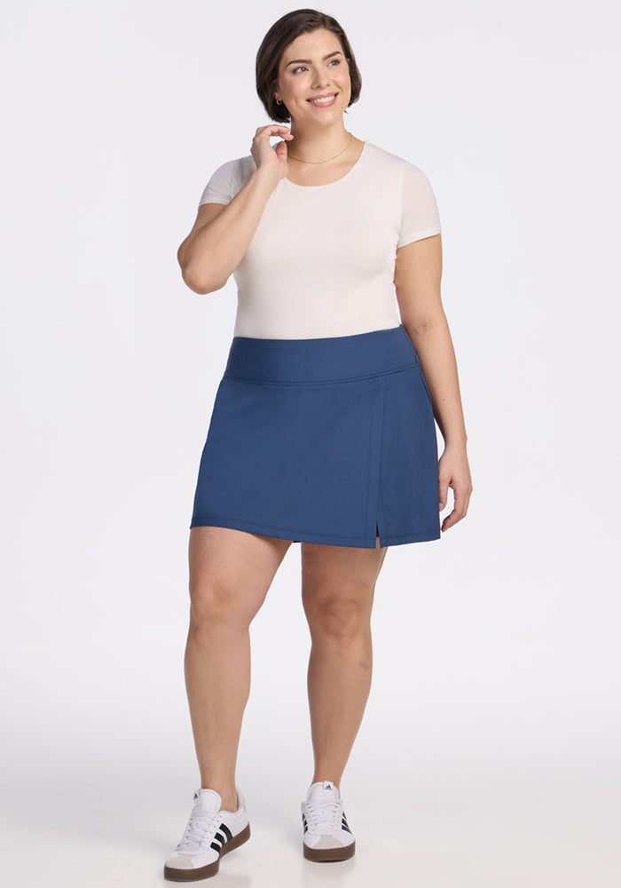 A woman smiles in the Woolx Topanga Skort in Vintage Indigo, styled with a white short-sleeve top and black-and-white sneakers, standing against a plain white background.
