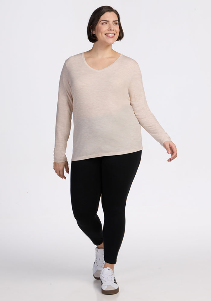 A woman with short dark hair smiles as she walks, wearing the Woolx Millie Long Sleeve - Cream Heather, black leggings, and white sneakers against a plain white background.