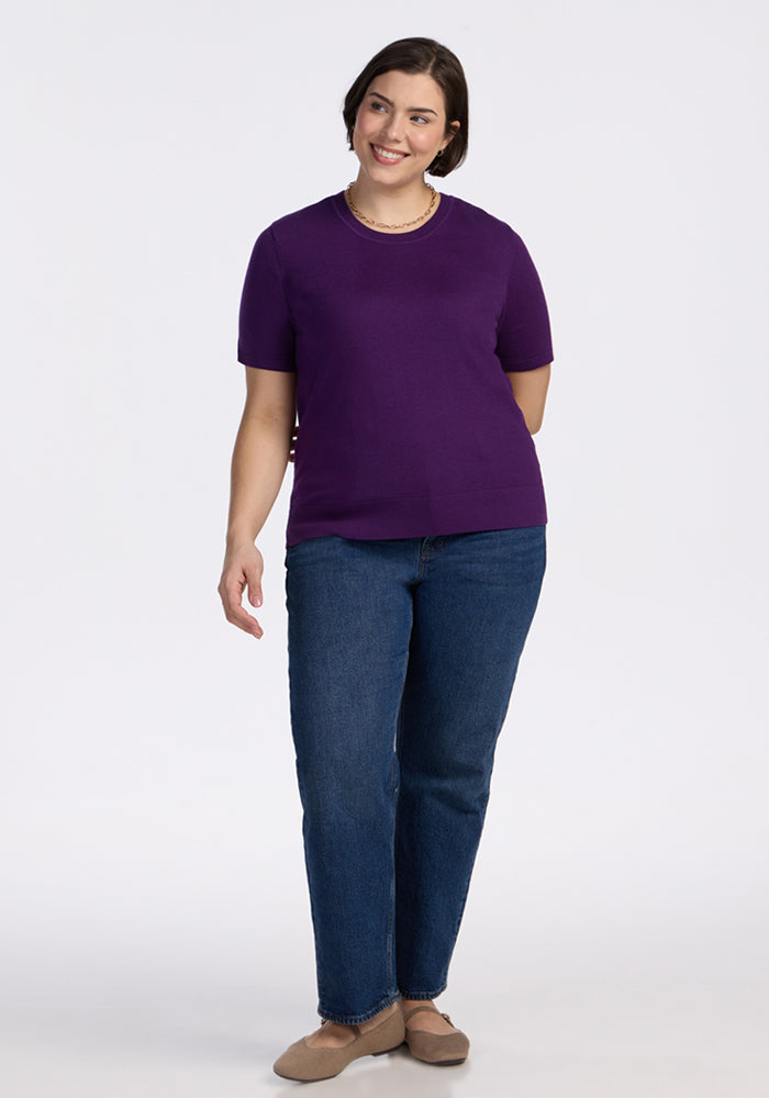 A woman with short brown hair smiles against a plain white background, wearing the Woolx Anna Crew Sweater in Plum Crush, blue jeans, beige shoes, and a pearl necklace.