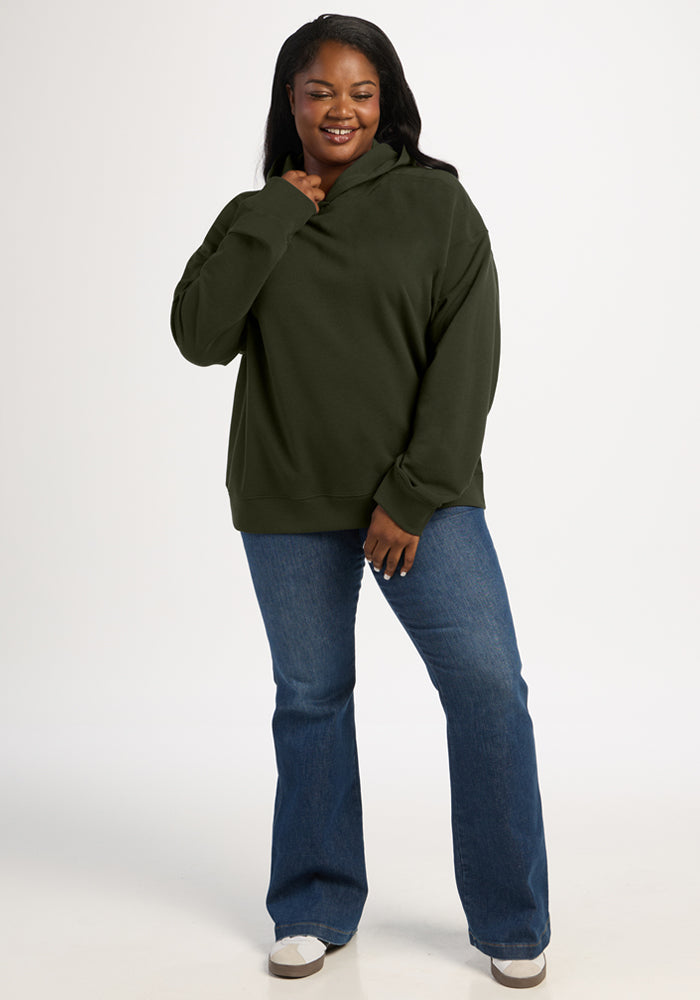 A smiling woman wearing a dark green Woolx Genevieve Hoodie - Duffel Bag and blue jeans.