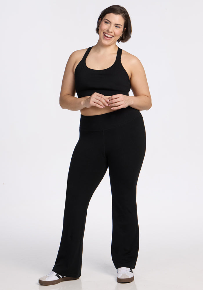 A woman with short brown hair smiles in a relaxed pose, wearing Woolx’s Morgan Flare Pants in black, a black sports bra, and white sneakers against a plain white background.