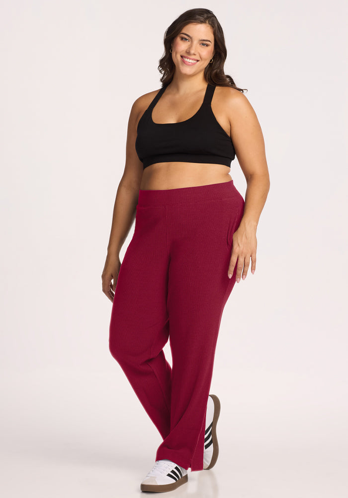 Smiling woman in Luca Ribbed Pants - Beet Red by Woolx, black sports bra, and sneakers poses against white background.