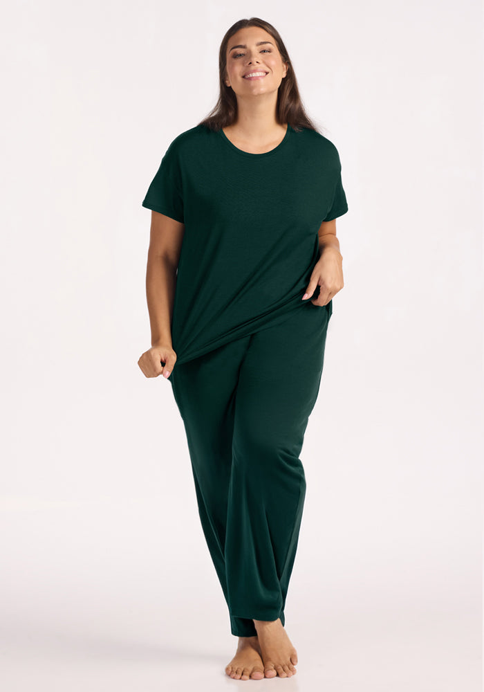 A smiling woman wears a dark green Billie Lounge Tee in Deep Pine and matching wide-leg merino wool pajama pants.