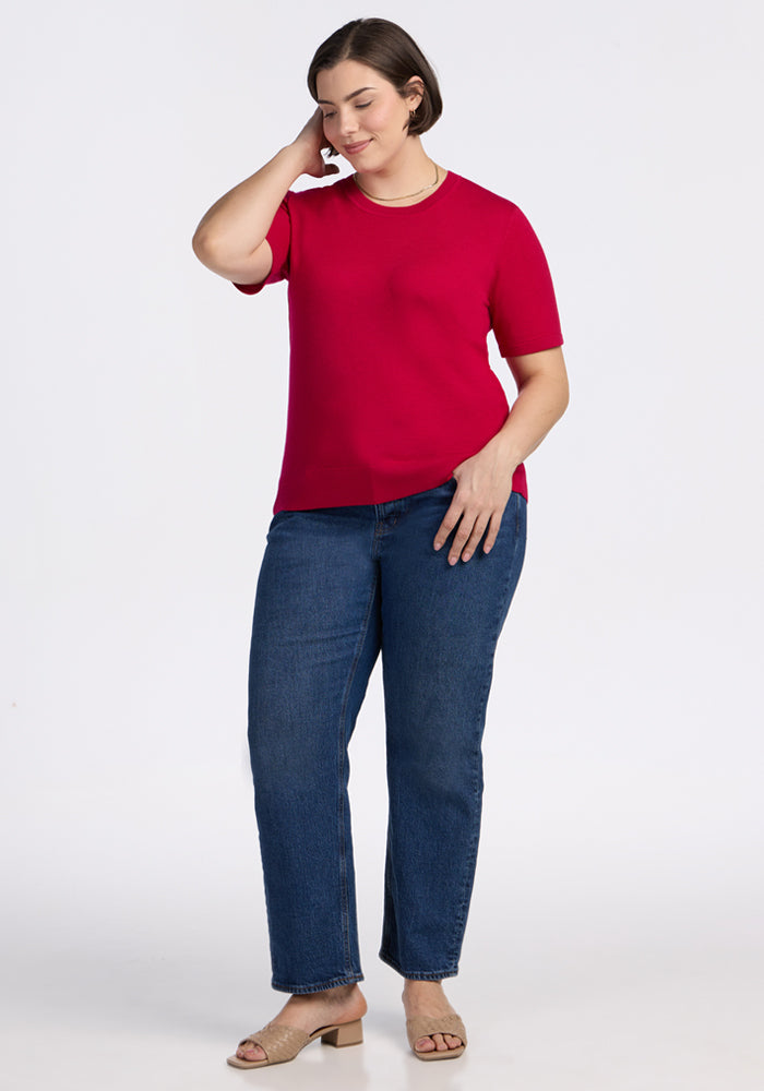 A woman stands against a plain white background, wearing the Woolx Anna Crew Sweater in Poppy Spark with blue jeans and beige heeled sandals. She touches her short brown hair and smiles softly.