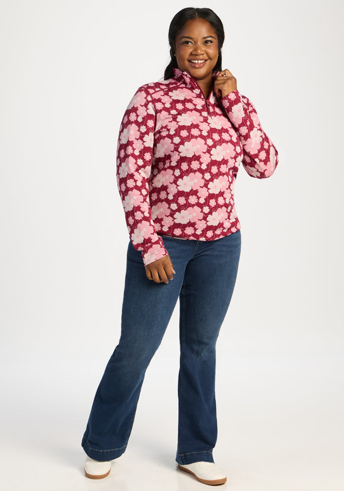 Smiling woman in Woolx Emi 1/4 Zip - Floral Crush, blue jeans, and white shoes stands on a plain white background.