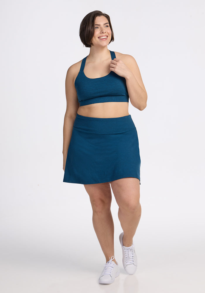 A woman with short brown hair smiles, wearing the Woolx Livi Bra in Majolica Blue and an athletic skirt. She touches her top with one hand, paired with white sneakers, all set against a plain white background.