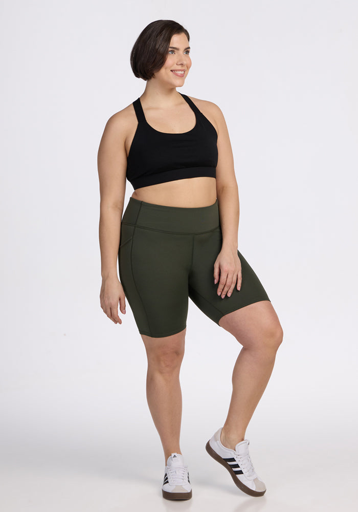 A woman in a black sports bra, Woolx Merino Flex® Dani Bike Shorts in Everleaf, and white sneakers smiles against a plain white background.
