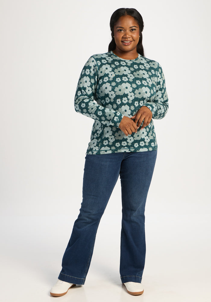 Woman wears Woolx Maddie Baselayer Top - Coastal Wildflower and blue jeans, smiling before a plain white background.
