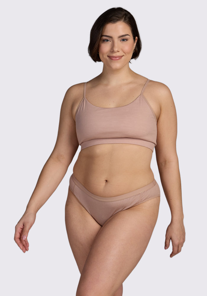 A woman with short brown hair smiles, wearing a nude bralette and Woolx Roxie Bikini Underwear in Morning Latte, standing against a plain light background.