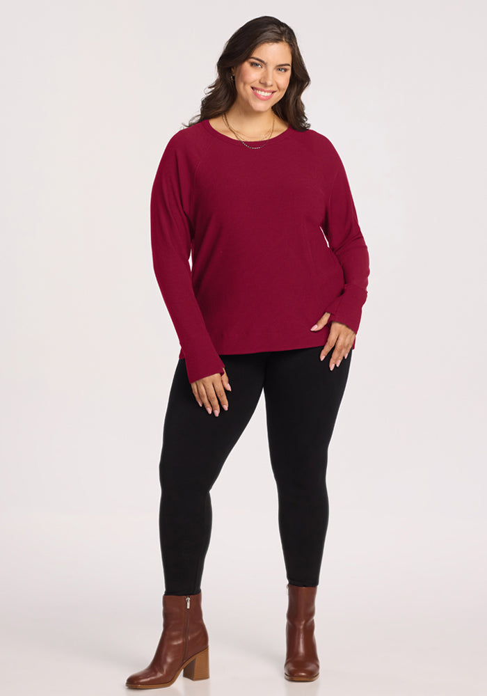 Smiling woman in Woolx Ainsley Sweater - Beet Red, black leggings, and brown boots stands against a white background.