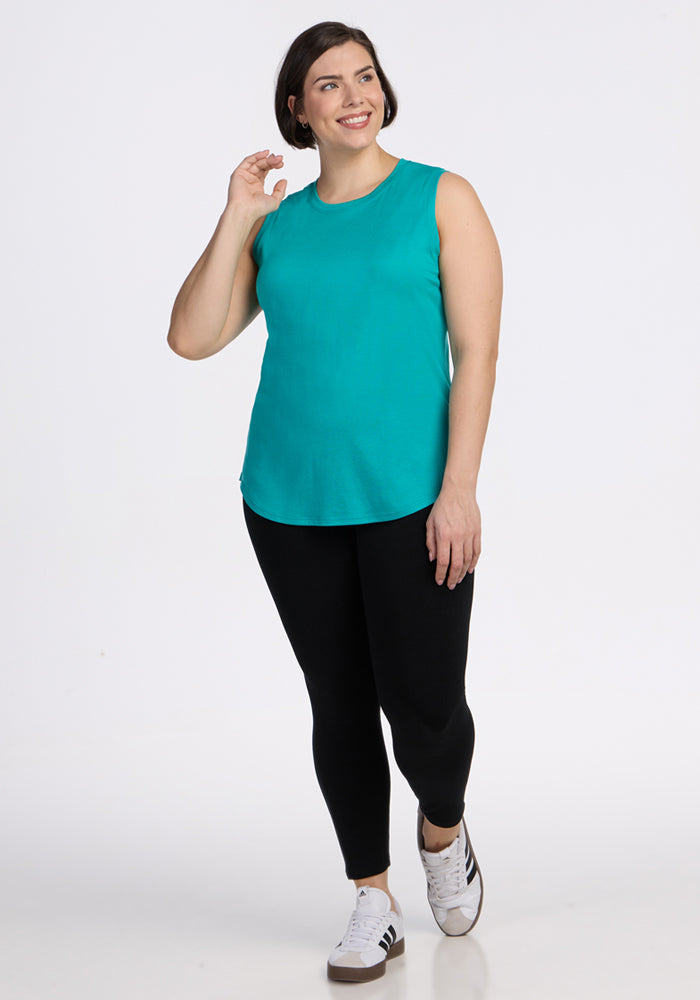 A woman smiles in a sleeveless Woolx Bella Tunic Top in Cool Jade, black leggings, and white striped sneakers, set against a plain white background.