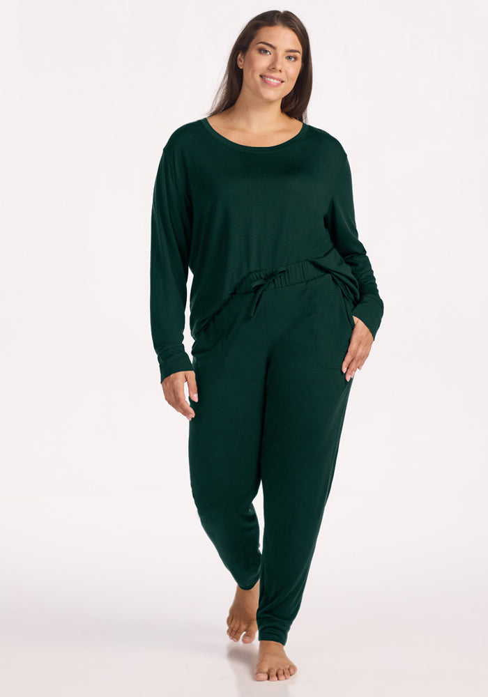 A woman wears a dark green long-sleeved shirt and matching Woolx Mary Kate Lounge Pants - Deep Pine joggers.