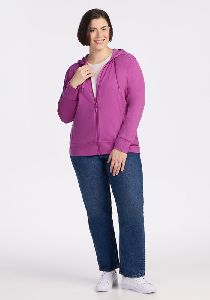 A woman with short brown hair, wearing the Woolx Ryann Hoodie in Purple Wine—her travel essential—over a white t-shirt, blue jeans, and white sneakers stands smiling, holding the hoodie zipper against a plain white background.