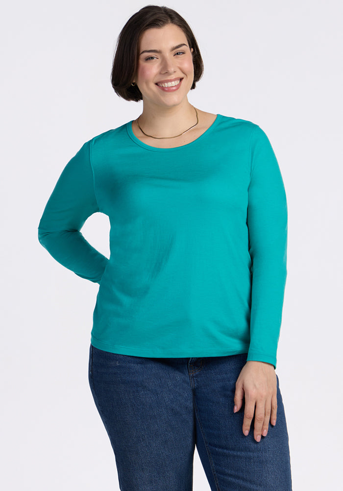 A person with short dark hair, wearing the Woolx Remi Long Sleeve T-Shirt in Cool Jade and blue jeans, stands smiling with one hand on their hip against a plain white background.